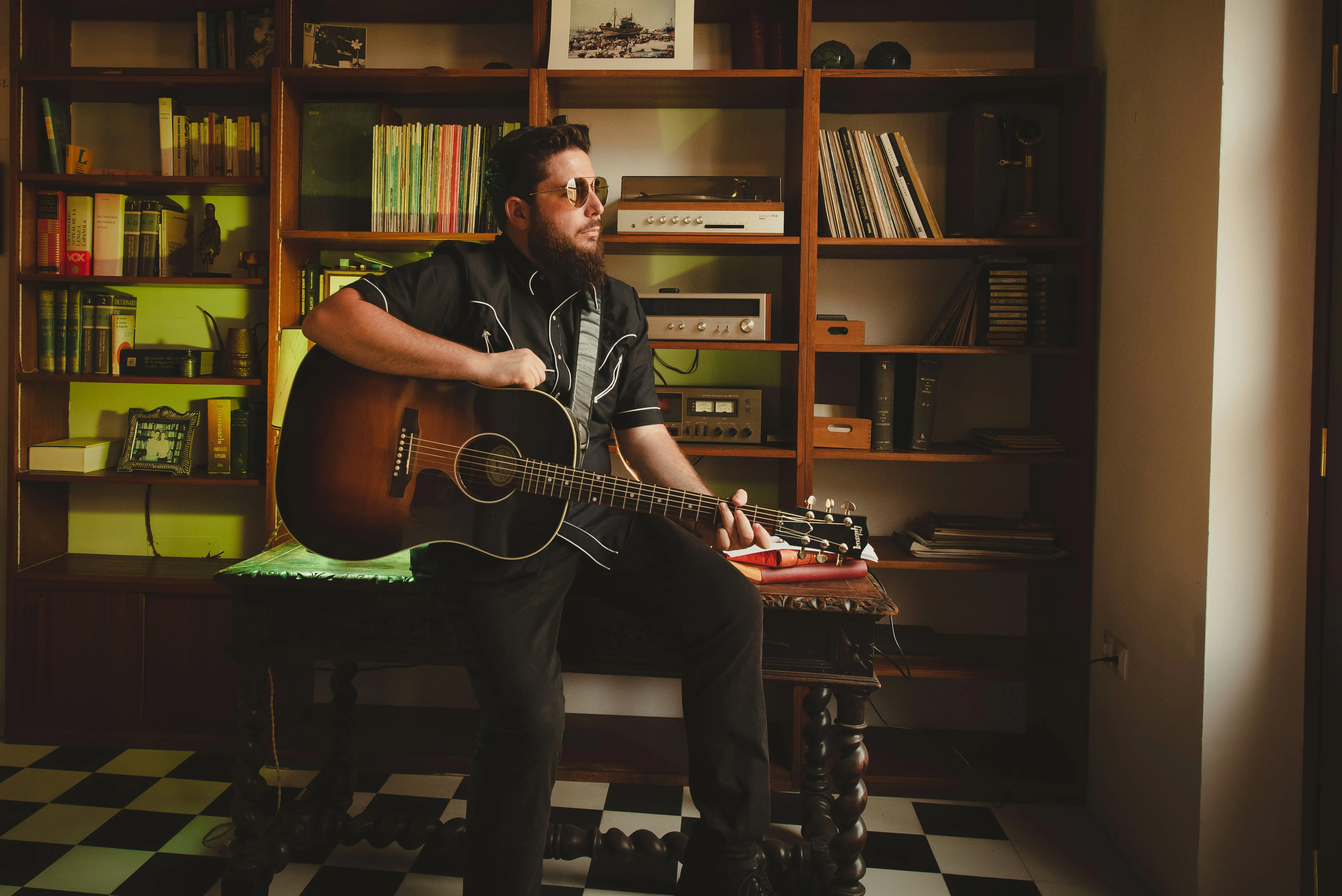 JJ Fuentes con una guitarra acústica, sentado ante una estantería de libros y vinilos