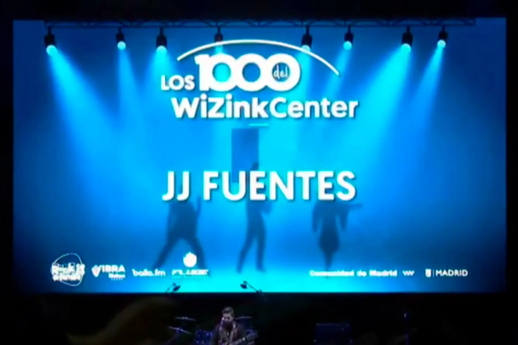 La pantalla gigante del WiZink Center con el rótulo «Los 1000 del WiZink Center» y debajo el nombre JJ Fuentes, con la banda diminuta sobre el escenario